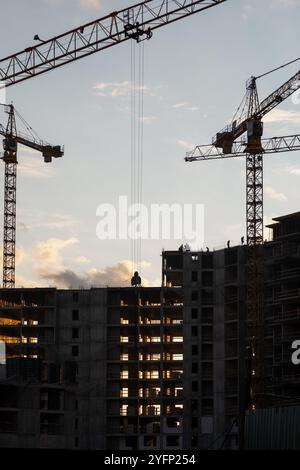 Le gru operano in un cantiere durante il tramonto, con i lavoratori visibili sulla struttura dell'edificio quando la luce del giorno svanisce Foto Stock