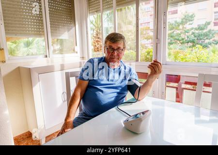 Uomo anziano in sovrappeso che misura la pressione sanguigna con il monitor a casa in soggiorno Foto Stock