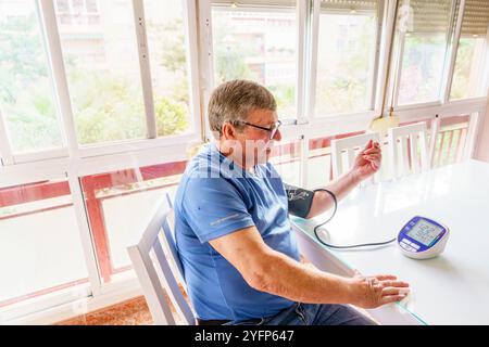 Uomo anziano in sovrappeso che misura la pressione sanguigna con il monitor a casa in soggiorno Foto Stock