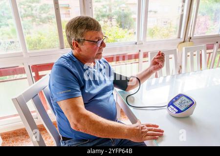 Uomo anziano in sovrappeso che misura la pressione sanguigna con il monitor a casa in soggiorno Foto Stock