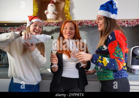 Donne d'affari sorridenti che indossano cappelli natalizi versando flute di champagne per celebrare il natale insieme in una cucina dell'ufficio Foto Stock
