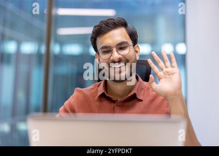 Allegro uomo d'affari ispanico che salta durante una videochiamata sul computer portatile in un ufficio luminoso. Comunicazione coinvolgente. Cattura l'interazione professionale gioiosa. Foto Stock