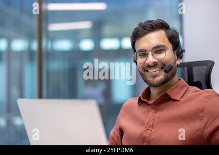 Uomo d'affari ispanico che indossa una cuffia che lavora su un computer portatile in ufficio. Sorriso sicuro, camicia marrone, occhiali, coinvolgente con professionalità. Sfondo luminoso e moderno. Affari e comunicazione. Foto Stock