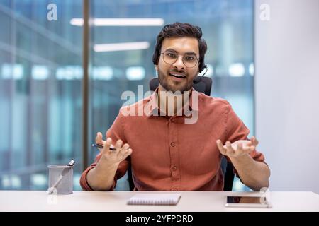 Uomo d'affari ispanico con cuffie in ufficio, impegnato in una conversazione sicura. Gli occhiali e gli abiti da ufficio migliorano l'immagine professionale, sottolineando la comunicazione e l'uso della tecnologia moderna. Foto Stock