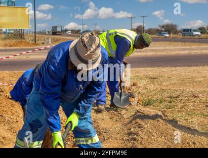 Due operai africani che scavano una trincea sul lato dell'autostrada installando fibre ottiche per l'azienda di telecomunicazioni Foto Stock