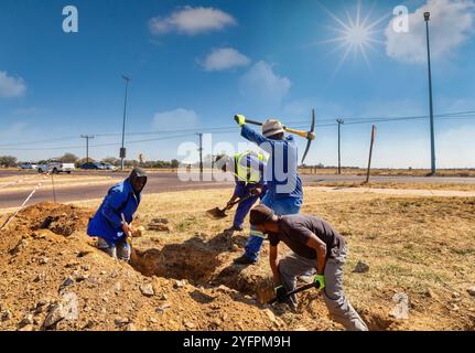 Gruppo di operai africani che scavano una trincea sul lato dell'autostrada installando fibre ottiche per l'azienda di telecomunicazioni Foto Stock