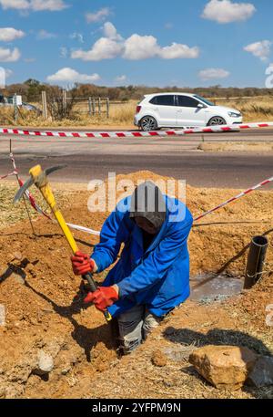 Lavoratore africano con un piccone che scava una trincea sul lato dell'autostrada riparando un tubo dell'acqua rotto nel fosso Foto Stock