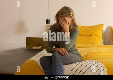 donna anziana seduta sul letto, sentendosi stressata e coprendo il viso con la mano, a casa Foto Stock