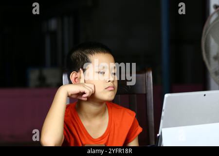 Un bambino si siede con cura a un tavolo, impegnato con un tablet in una stanza poco illuminata, mostrando la miscela di curiosità e concentrazione nell'apprendimento digitale Foto Stock