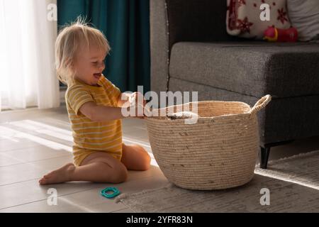 Un bambino piccolo con un onesie a strisce gialle si inginocchia giocando con un gatto che riposa all'interno di un comodo cesto in tessuto. La luce del sole scorre attraverso un vicino Foto Stock