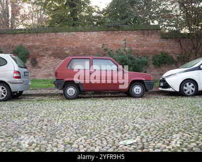 Cremona, Lombardia, Italia 27 ottobre 2024 piccolo panda rosso fiat parcheggiato in una piazza acciottolata accanto ad un muro di mattoni, con una moderna auto bianca parcheggiata accanto Foto Stock