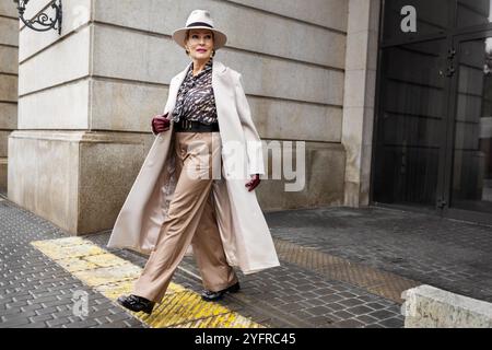 Elegante donna matura che indossa abbigliamento alla moda, cappotto e pantaloni beige, cappello in feltro, accessori, cintura e guanti in pelle cammina contro la parete beige in C. Foto Stock