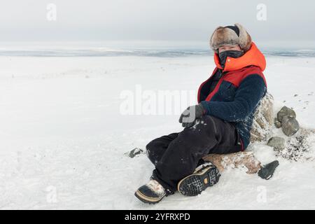 Musher maschile caucasico adulto vestito con attrezzatura artica su un paesaggio innevato di tundra, di fronte a freddo estremo, in una natura selvaggia ghiacciata, slitte trainate da cani in bac Foto Stock