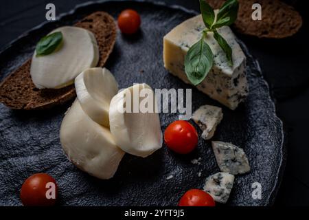 Mozzarella e garganzola blu con pane di segale scuro e pomodori ciliegini con basilico verde su un piatto di mica nera su sfondo scuro. Foto di alta qualità Foto Stock