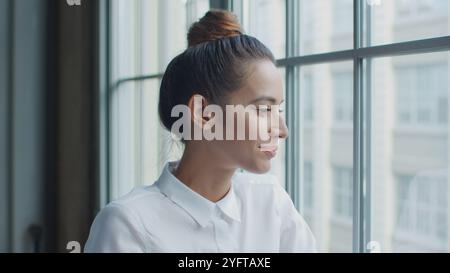 Testa e spalle Ritratto di una giovane donna d'affari sorridente che guarda fuori dalla finestra in ufficio Foto Stock