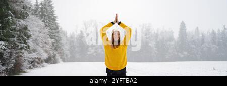 Una donna che indossa un maglione giallo e un cappello bianco tiene una posa di yoga in una serena radura coperta di neve, circondata da alberi ghiacciati sotto un morbido e coperto Foto Stock