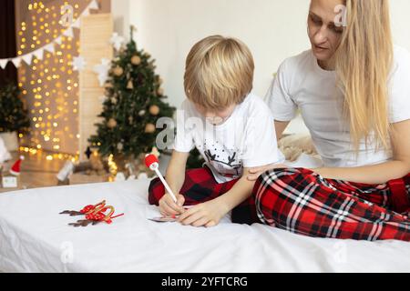 Mamma e figlio di 7 anni in pigiama natalizio, seduti a letto, scrivi una lettera a Babbo Natale o cartolina per gli amici, sognando regali. Buon Christm Foto Stock