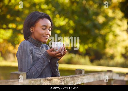 Vista del profilo di una giovane donna a piedi nella campagna autunnale appoggiata su Fence che tiene una bevanda calda Foto Stock