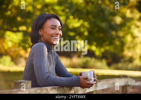 Vista del profilo di una giovane donna a piedi nella campagna autunnale appoggiata su Fence che tiene una bevanda calda Foto Stock