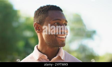 Un uomo allegro sta passeggiando attraverso un parco vivace, crogiolandosi alla luce calda del sole. Il suo sorriso luminoso riflette la gioia del momento mentre gli piace il bea Foto Stock