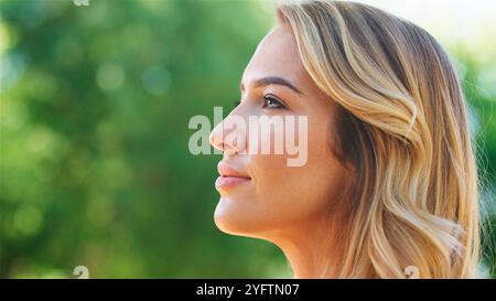 Una giovane donna dai capelli ondulati guarda attentamente in lontananza, il suo profilo illuminato dalla luce del sole. Si trova fuori in un lussureggiante parco verde, Surro Foto Stock