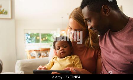 Una famiglia amorevole si siede da vicino su un divano, impegnata a guardare contenuti su un tablet. I genitori condividono un momento di gioia con il loro bambino, che appare Foto Stock