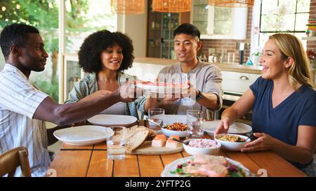 Un gruppo di quattro amici condivide un momento di gioia al tavolo da pranzo, passando per i piatti pieni di cibo. Risate e conversazione riempiono l'aria come Foto Stock