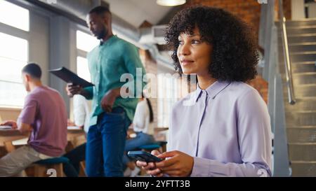 Donna d'affari sorridente che utilizza il telefono cellulare che lavora in Open Plan Office con colleghi in background Foto Stock