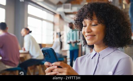 Donna d'affari sorridente che utilizza il telefono cellulare che lavora in Open Plan Office con colleghi in background Foto Stock