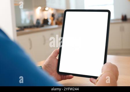 Una persona è seduta a un tavolo da cucina, tenendo in mano un tablet vuoto mentre guarda il suo schermo. La cucina presenta un design aperto e arioso Foto Stock