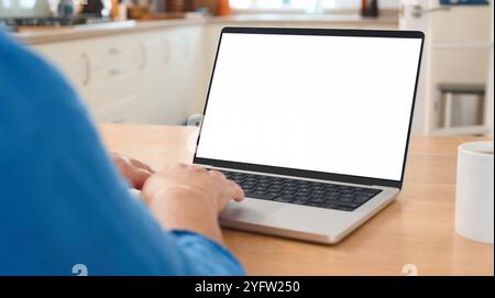 Una persona si concentra sulla digitazione su un notebook posizionato su un tavolo di legno in una cucina moderna. L'ambiente luminoso e i toni caldi suggeriscono un intimo Foto Stock
