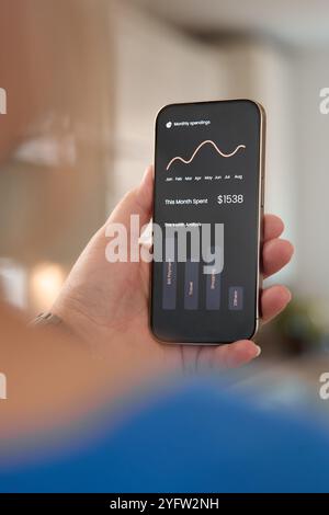 Una persona sta esaminando il proprio dashboard finanziario su uno smartphone, mostrando i guadagni e le tendenze mensili. L'ambiente sembra essere un ambiente accogliente Foto Stock