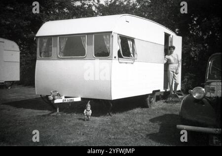 1950, storica, una donna in piedi su un gradino di metallo all'ingresso della sua carovana delle vacanze, un'auto dell'epoca parcheggiata accanto....piccolo gnomo da giardino sull'erba, Inghilterra, Regno Unito. Foto Stock