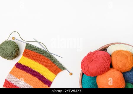 Tavolo da lavoro con abiti caldi per maglieria. Palline di filo arancione, rosso, verde e giallo nel cestello, sciarpa a righe multicolore e aghi per maglieria. Concetto di ricamo Foto Stock