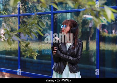 Immagine di una ragazza con un abito blu, giacca in pelle nera, con occhiali blu che guardano lontano contro una parete di vetro blu. Foto Stock