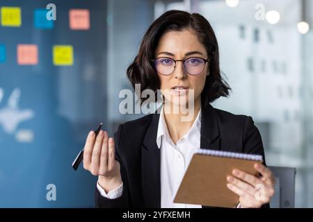 Giovane donna d'affari in ufficio che discute idee, indossa occhiali, tiene in mano il blocco note e la penna. Leader donna sicura di sé si impegna in riunioni professionali, comunica strategie, guida le discussioni di squadra. Foto Stock