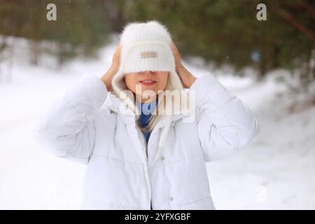 una donna che indossa un comodo cappello in maglia bianca e un cappotto bianco coordinato, in piedi in mezzo a una foresta serena ricoperta di neve fresca. Con uno sguardo di gioia e meraviglia sul viso, abbraccia pienamente la bellezza dell'inverno e la magia della prima nevicata Foto Stock