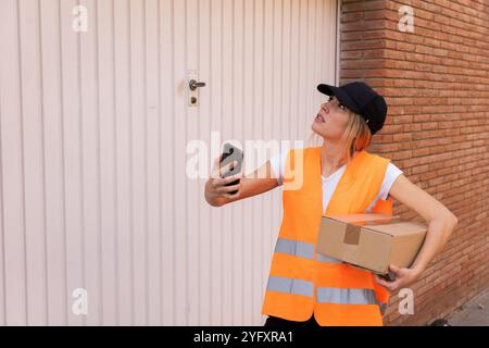 Una donna che lavora come corriere sembra perduta. Tiene il telefono cellulare in una mano e con l'altra una scatola di cartone da consegnare Foto Stock