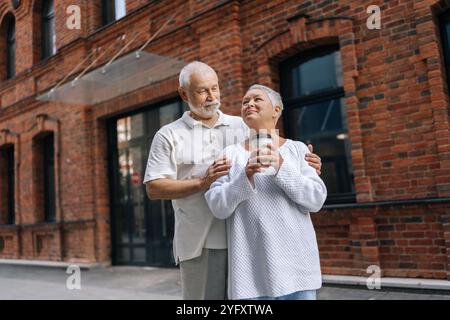 Affascinante uomo e donna anziana in piedi su strada asfaltata urbana, tenendo tazze di caffè e amando guardarsi l'un l'altro. Felice coppia senior che si diverte Foto Stock