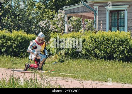 Il cavaliere medievale in piena armatura si inginocchia a terra nel campo dell'erba. Indossa casco e tuta in metallo e fissa Foto Stock