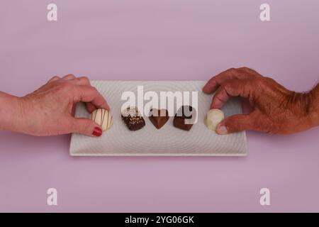 Mani che selezionano cioccolatini assortiti su vassoio bianco su sfondo rosa Foto Stock