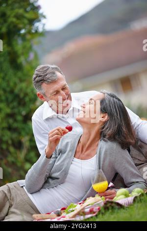 Maturo, coppia e mangiare al picnic all'aperto con frutta sana, Unione e cestino di cibo di data romantica. Gente, ridere e amare con il succo Foto Stock