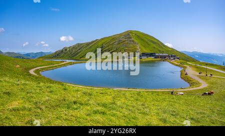 Il serbatoio d'acqua Wildkogel riflette il cielo azzurro, circondato da lussureggianti colline verdi. I visitatori apprezzano il tranquillo paesaggio mentre esplorano i sentieri e ammirano le splendide viste alpine. Foto Stock