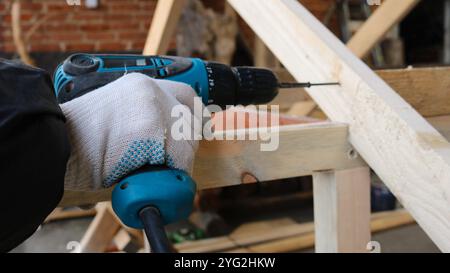 Un uomo che indossa abiti da lavoro e guanti fissa un cacciavite in una struttura di legno contro una parete di mattoni, assemblando una struttura da blocchi di legno Foto Stock