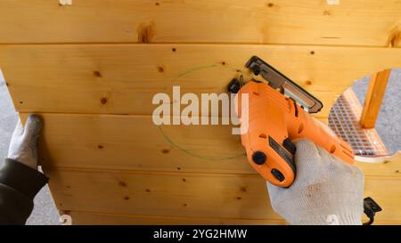 Un lavoratore con i guanti utilizza un seghetto elettrico per vedere un cerchio fuori da una parete di legno in base ai contrassegni. Foto Stock