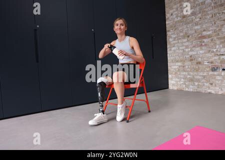 Donna con una gamba protesica seduta su una sedia e con una bottiglia d'acqua in palestra Foto Stock