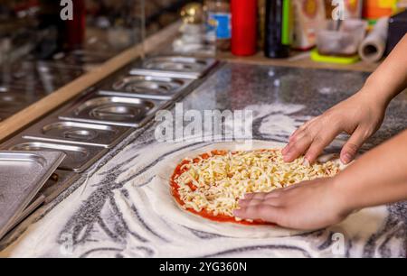 Uomo che aggiunge formaggio alla pizza a tavola, primo piano. Concetto di cucina: Cuocere a mano aggiungendo formaggio grattugiato alla pizza alla pizzeria. Cuocere in cucina mettendo il Foto Stock