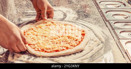 Primo piano della mano dello chef baker che prepara la pizza in cucina. Uomo che aggiunge formaggio alla pizza a tavola, primo piano. Concetto di cottura, cuocere a mano aggiungendo formaggio grattugiato a. Foto Stock