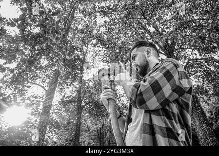 Falegname hipster con un'ascia. Taglierini barbuti in camicia a scacchi tagliando il tronco alla segheria. Lumberjack. Tritare la legna. Bianco e nero Foto Stock