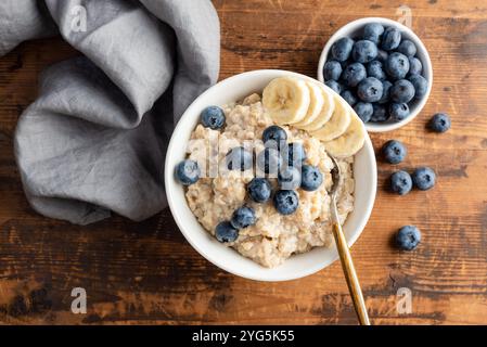 Sana farina d'avena condita con mirtilli freschi e fette di banana, servita in una ciotola bianca su un tavolo di legno. Vista dall'alto Foto Stock